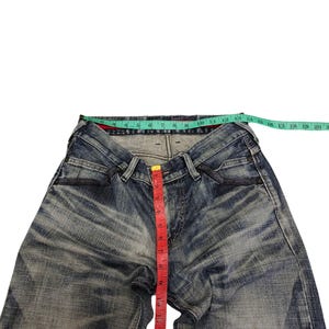 May include: Blue denim jeans with a faded wash, featuring a red measuring tape indicating a length of approximately 14 inches. A green measuring tape shows the waist size. The jeans have a classic five-pocket design.