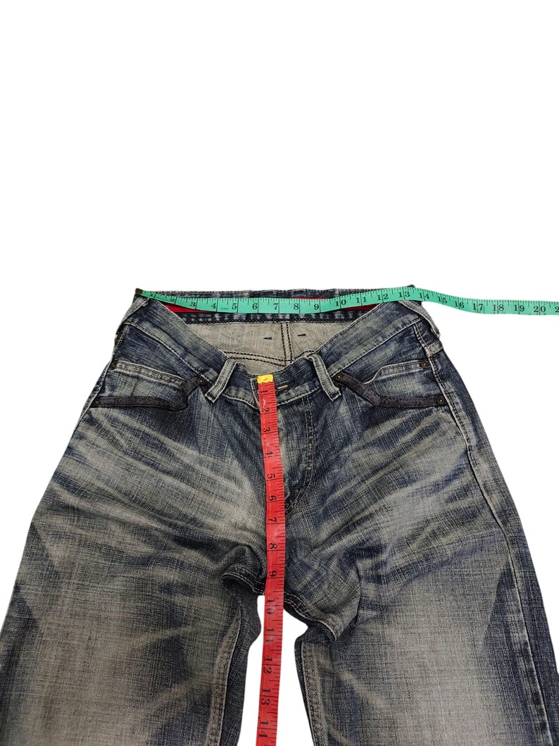 May include: Blue denim jeans with a faded wash, featuring a red measuring tape indicating a length of approximately 14 inches. A green measuring tape shows the waist size. The jeans have a classic five-pocket design.