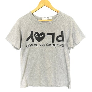 Może przedstawiać: Szary T-shirt z czarnym logo Play Comme des Gar&ccedil;ons przedstawiającym serce z oczami. Logo jest nadrukowane w odwr&oacute;conej czcionce.