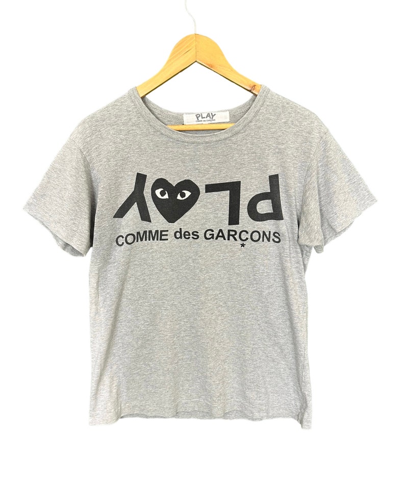 Może przedstawiać: Szary T-shirt z czarnym logo Play Comme des Gar&ccedil;ons przedstawiającym serce z oczami. Logo jest nadrukowane w odwr&oacute;conej czcionce.