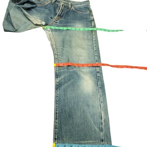 Puede incluir: Vaqueros de mezclilla azul claro con un lavado descolorido. Los vaqueros se miden con una cinta m&eacute;trica verde, naranja y azul. Los vaqueros est&aacute;n colocados sobre una superficie blanca. Los vaqueros tienen un dise&ntilde;o cl&aacute;sico de cinco bolsillos.