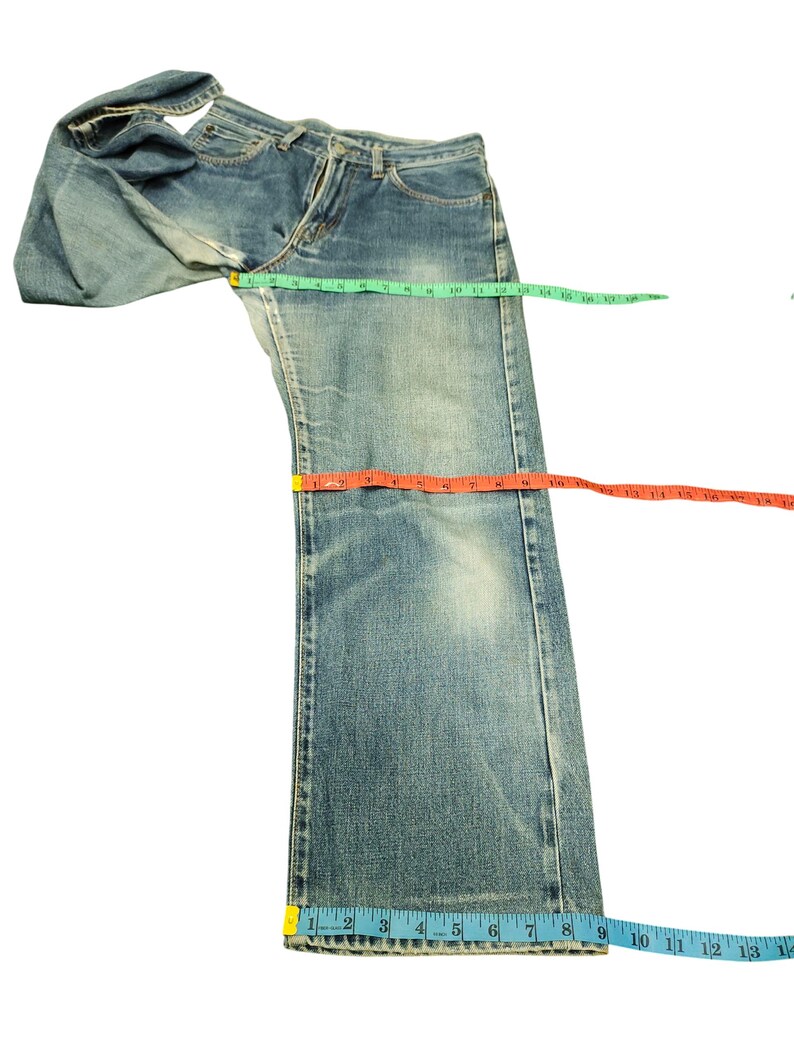 Puede incluir: Vaqueros de mezclilla azul claro con un lavado descolorido. Los vaqueros se miden con una cinta m&eacute;trica verde, naranja y azul. Los vaqueros est&aacute;n colocados sobre una superficie blanca. Los vaqueros tienen un dise&ntilde;o cl&aacute;sico de cinco bolsillos.