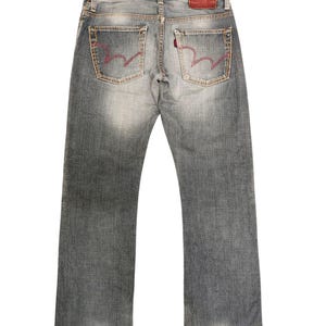 May include: Faded gray denim jeans with red stitching on the back pockets. The jeans have a classic five-pocket design, a red leather patch, and a straight leg cut. The jeans are a medium wash with subtle fading.