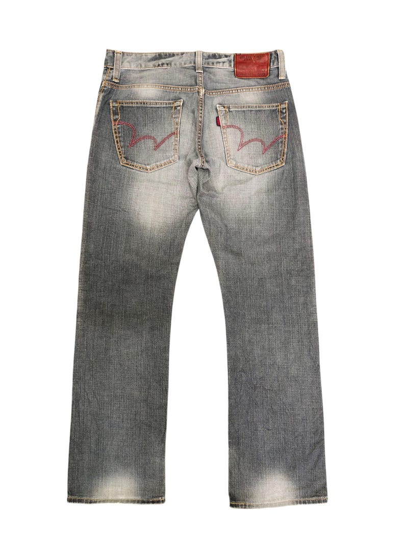 May include: Faded gray denim jeans with red stitching on the back pockets. The jeans have a classic five-pocket design, a red leather patch, and a straight leg cut. The jeans are a medium wash with subtle fading.
