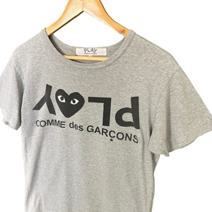 Może przedstawiać: Szary T-shirt z czarno-białym nadrukiem graficznym serca z oczami i tekstem "PLAY COMME des GARCONS*"