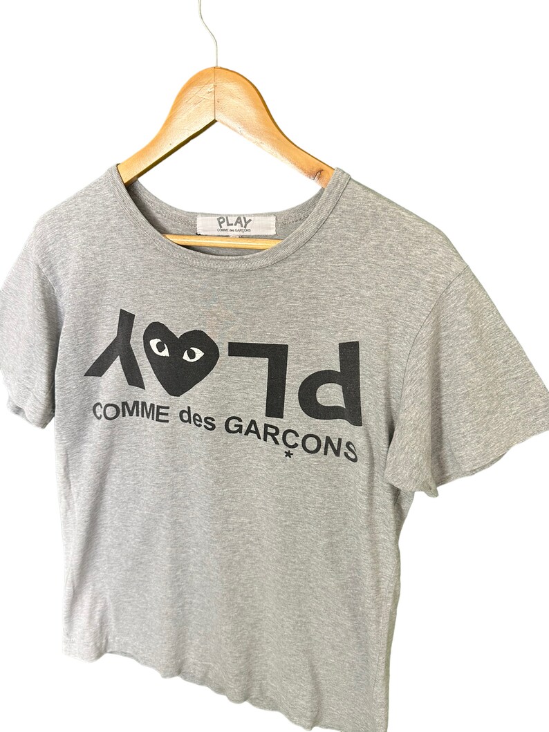 Może przedstawiać: Szary T-shirt z czarno-białym nadrukiem graficznym serca z oczami i tekstem "PLAY COMME des GARCONS*"