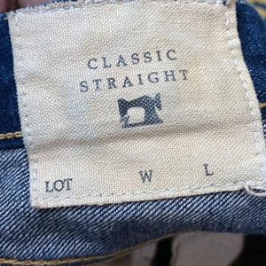 以下が含まれることがあります： デニムジーンズのラベルのクローズアップ。ラベルはベージュで、大文字で「CLASSIC STRAIGHT」と書かれています。中央にはミシンのアイコンがあります。テキストの下には「LOT」、「W」、「L」の文字があります。