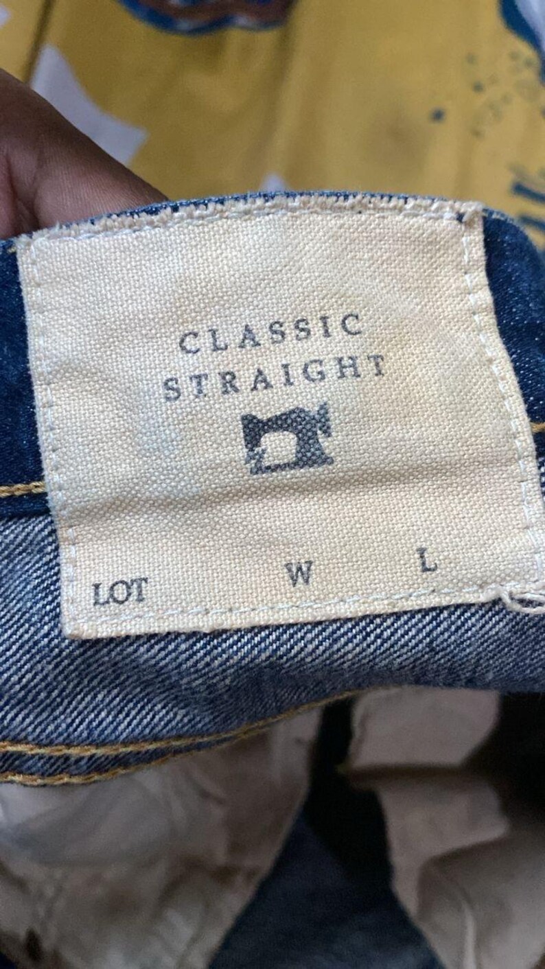 以下が含まれることがあります： デニムジーンズのラベルのクローズアップ。ラベルはベージュで、大文字で「CLASSIC STRAIGHT」と書かれています。中央にはミシンのアイコンがあります。テキストの下には「LOT」、「W」、「L」の文字があります。