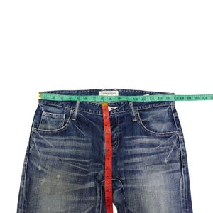 May include: Blue denim jeans with a distressed look, featuring a red measuring tape. The jeans have a button and zipper closure, pockets, and the brand name "VANQUISH" is visible. The waist measures approximately 12 inches.