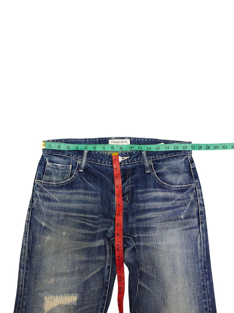 May include: Blue denim jeans with a distressed look, featuring a red measuring tape. The jeans have a button and zipper closure, pockets, and the brand name "VANQUISH" is visible. The waist measures approximately 12 inches.