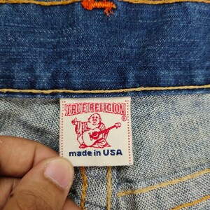 May include: Close-up of a blue denim jean with a red and white True Religion label that says "made in USA".