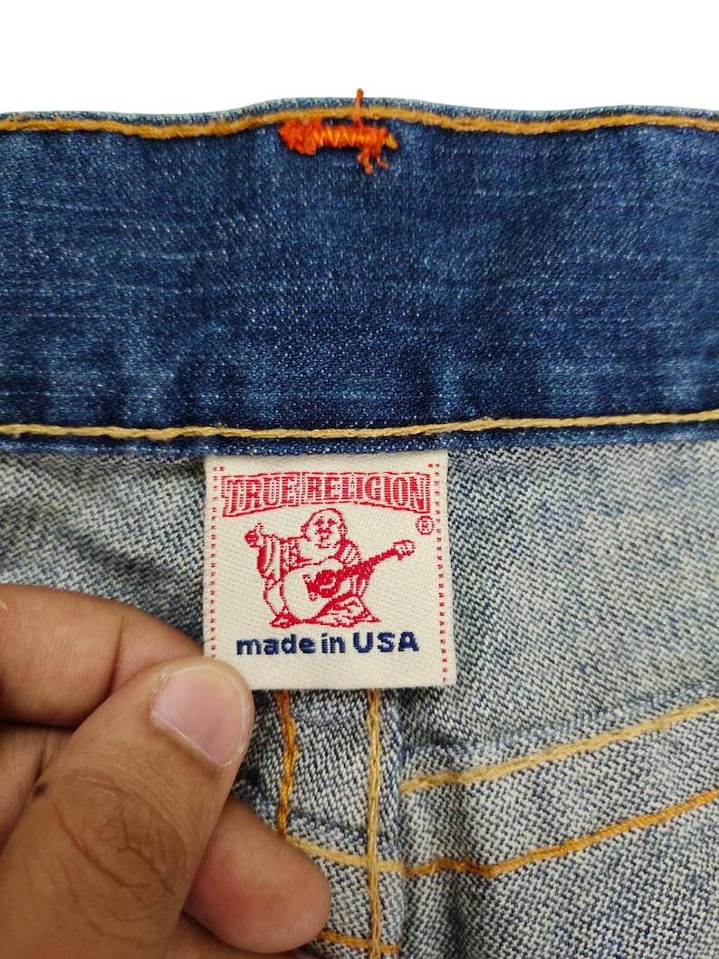 May include: Close-up of a blue denim jean with a red and white True Religion label that says "made in USA".