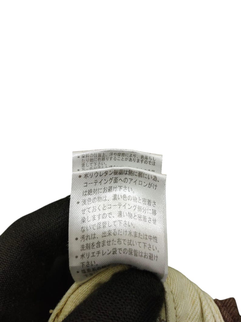 Puede incluir: Primer plano de una etiqueta de ropa con texto en japon&eacute;s. La etiqueta blanca con texto negro est&aacute; sujeta por una mano enguantada. La etiqueta proporciona instrucciones de cuidado para la prenda.