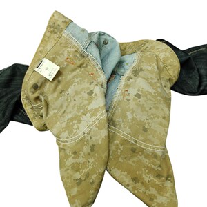 Op de afbeelding: Close-up van een camouflage-jeans met een lichtbruin en tan digitaal camopatroon. De jeans is opgevouwen, waardoor de lichtblauwe binnenkant en witte stiksels zichtbaar zijn. Een label met tekst is zichtbaar.