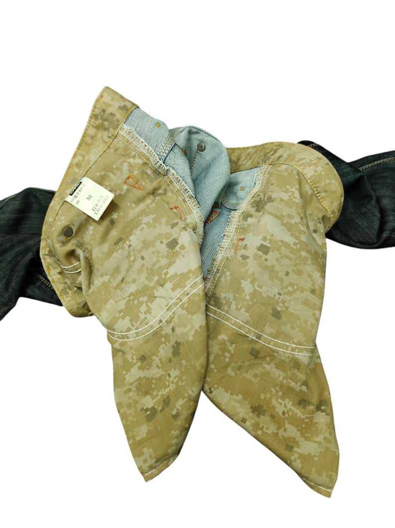 Op de afbeelding: Close-up van een camouflage-jeans met een lichtbruin en tan digitaal camopatroon. De jeans is opgevouwen, waardoor de lichtblauwe binnenkant en witte stiksels zichtbaar zijn. Een label met tekst is zichtbaar.