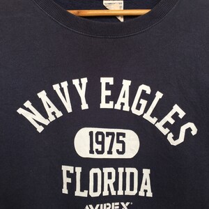 May include: A navy blue sweatshirt with white lettering that reads "NAVY EAGLES 1975 FLORIDA AVIREX".