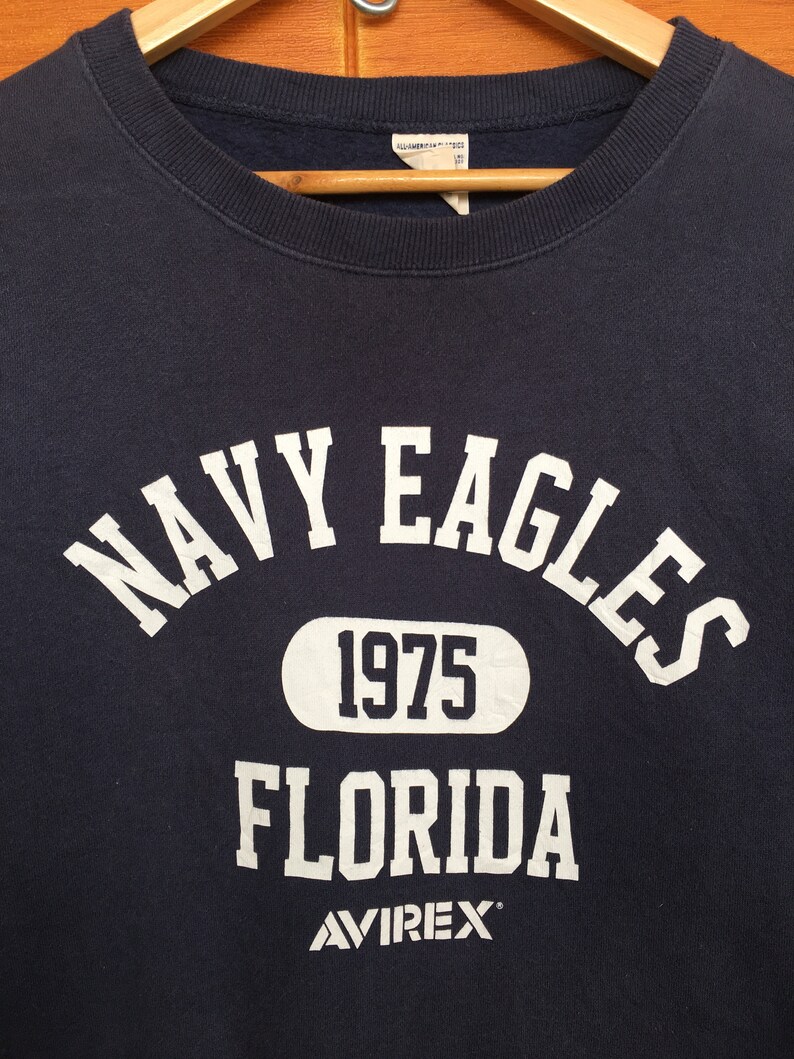 May include: A navy blue sweatshirt with white lettering that reads "NAVY EAGLES 1975 FLORIDA AVIREX".