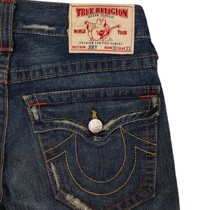 K&ouml;nnte beinhalten: Eine blaue Jeanshose mit einer Ges&auml;&szlig;tasche, die das True Religion-Markenlogo und den Text "World Tour Fashion for the Senses Section Joey Row 30 Seat 33" tr&auml;gt.