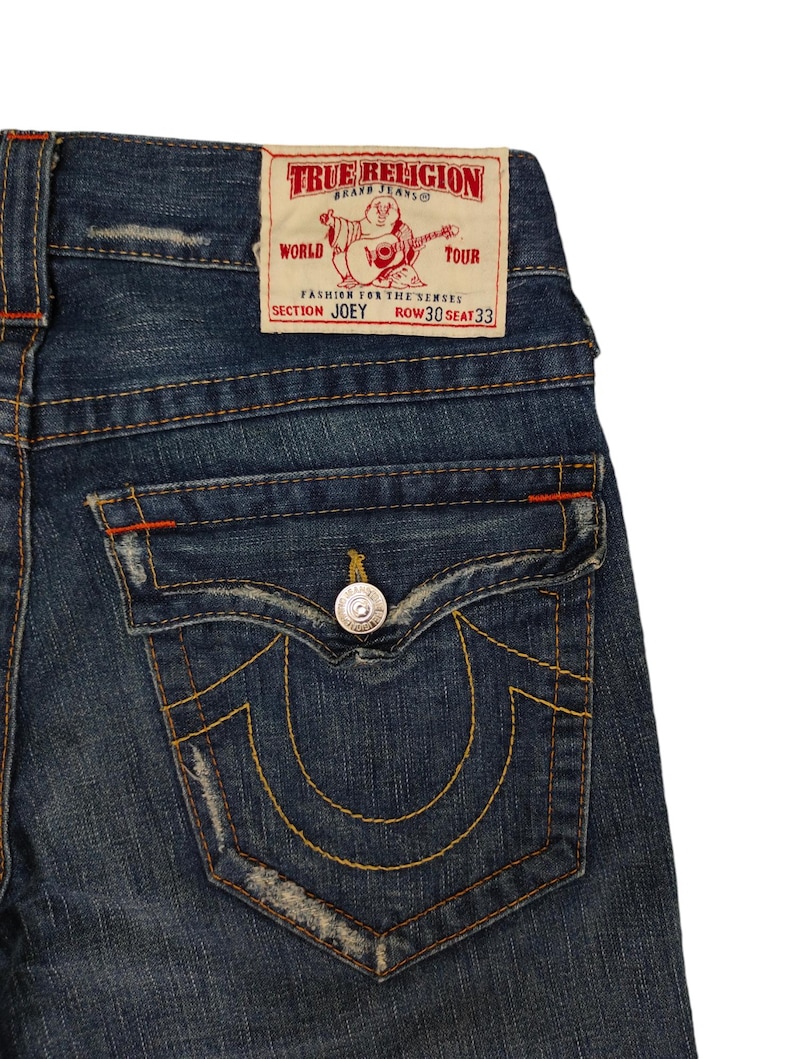 K&ouml;nnte beinhalten: Eine blaue Jeanshose mit einer Ges&auml;&szlig;tasche, die das True Religion-Markenlogo und den Text "World Tour Fashion for the Senses Section Joey Row 30 Seat 33" tr&auml;gt.