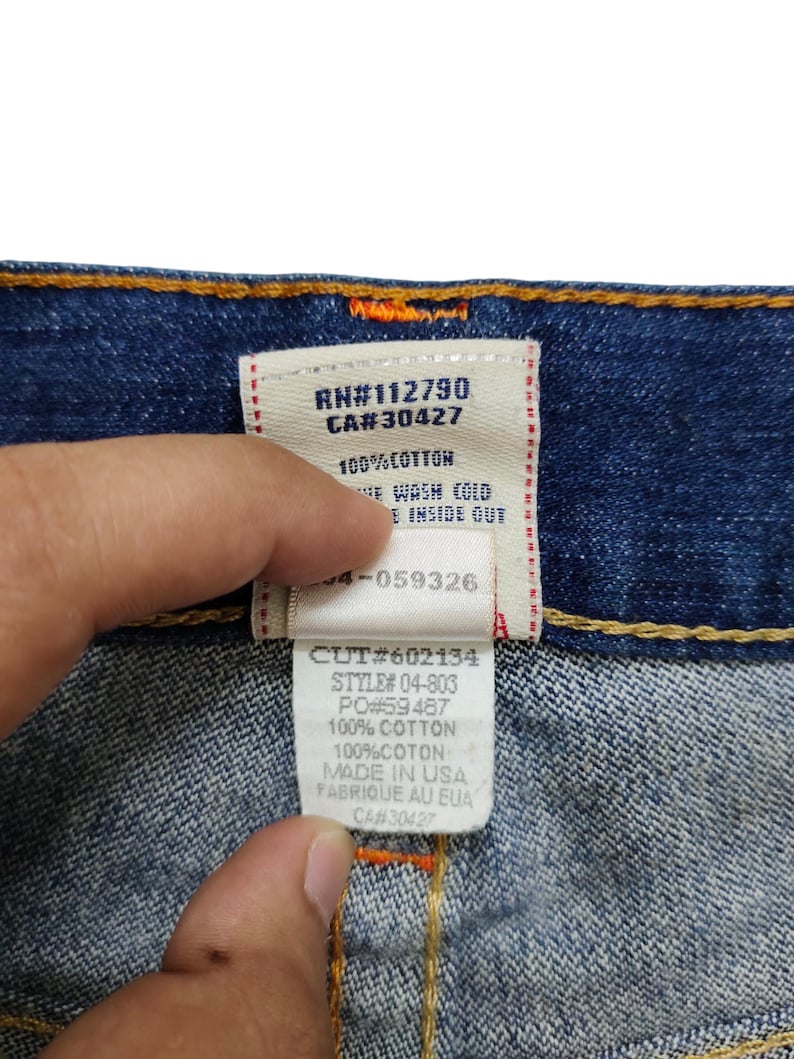 K&ouml;nnte beinhalten: Ein blaues Jeans-Etikett mit wei&szlig;em Text. Das Etikett lautet "RN#112790 CA#30427 100% COTTON NE WASH COLD INSIDE OUT 4-059326 CUT#602134 STYLE# 04-803 PO#59487 100% COTTON 100%COTON MADE IN USA FABRIQUE AU EUA CA#30427"