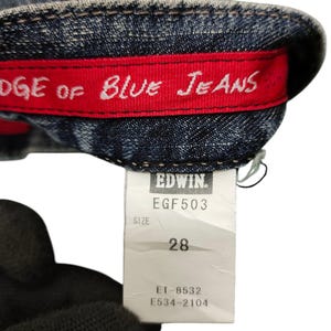 May include: Close-up of blue denim jeans with a red interior band that reads "EDGE OF BLUE JEANS" in white lettering. A white tag displays "EDWIN" and size 28.