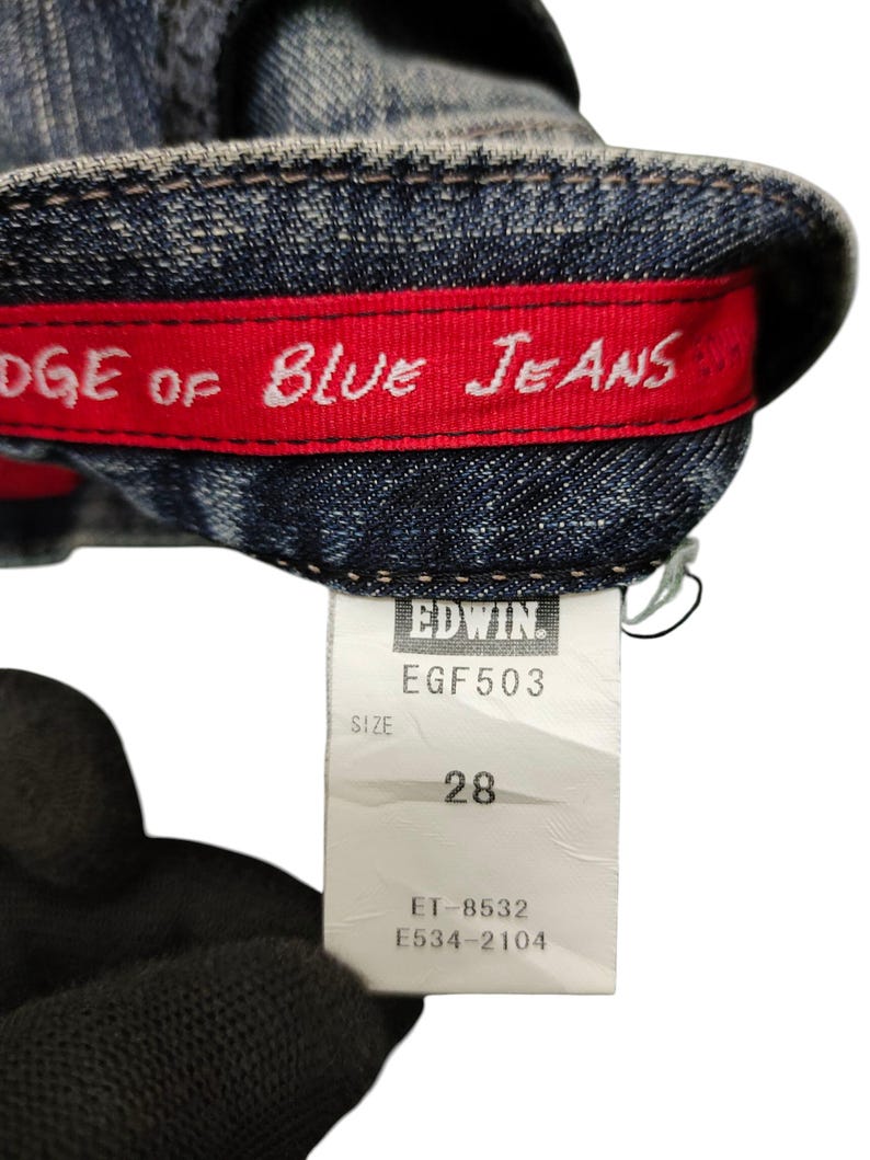 May include: Close-up of blue denim jeans with a red interior band that reads "EDGE OF BLUE JEANS" in white lettering. A white tag displays "EDWIN" and size 28.