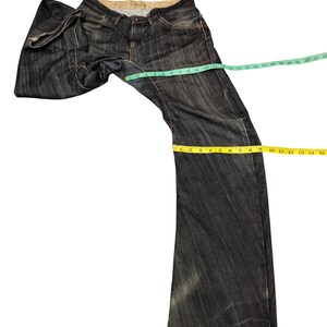 Op de afbeelding: Donkerblauwe denim jeans met een vervaagde uitstraling. De jeans worden gemeten met een groen, geel en blauw meetlint. De jeans hebben een klassiek ontwerp met vijf zakken en een knoop- en ritssluiting.