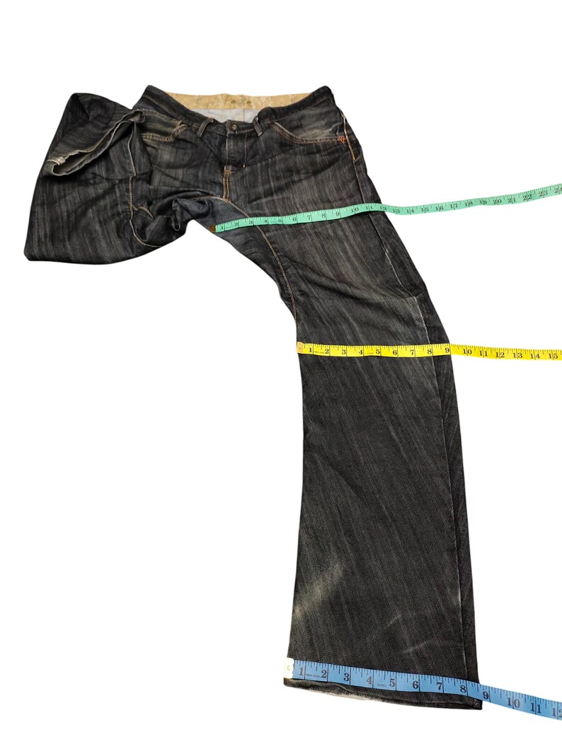 Op de afbeelding: Donkerblauwe denim jeans met een vervaagde uitstraling. De jeans worden gemeten met een groen, geel en blauw meetlint. De jeans hebben een klassiek ontwerp met vijf zakken en een knoop- en ritssluiting.