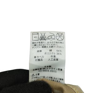 Op de afbeelding: Close-up van een kledinglabel met Japanse tekst en wassymbolen. Het label bevat details over de stof: 64% katoen, 36% polyester, met een leren label en accessoires van kunstleer. Wasvoorschriften en fabrikantgegevens zijn ook inbegrepen.