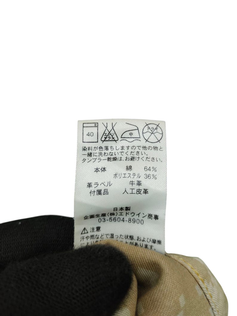 Op de afbeelding: Close-up van een kledinglabel met Japanse tekst en wassymbolen. Het label bevat details over de stof: 64% katoen, 36% polyester, met een leren label en accessoires van kunstleer. Wasvoorschriften en fabrikantgegevens zijn ook inbegrepen.