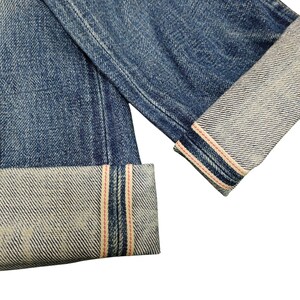 Puede incluir: Primer plano de vaqueros azules con pu&ntilde;os doblados. Los pu&ntilde;os revelan una tela m&aacute;s clara con rayas rojas y blancas a lo largo de los bordes. El denim tiene un aspecto desgastado y texturizado.