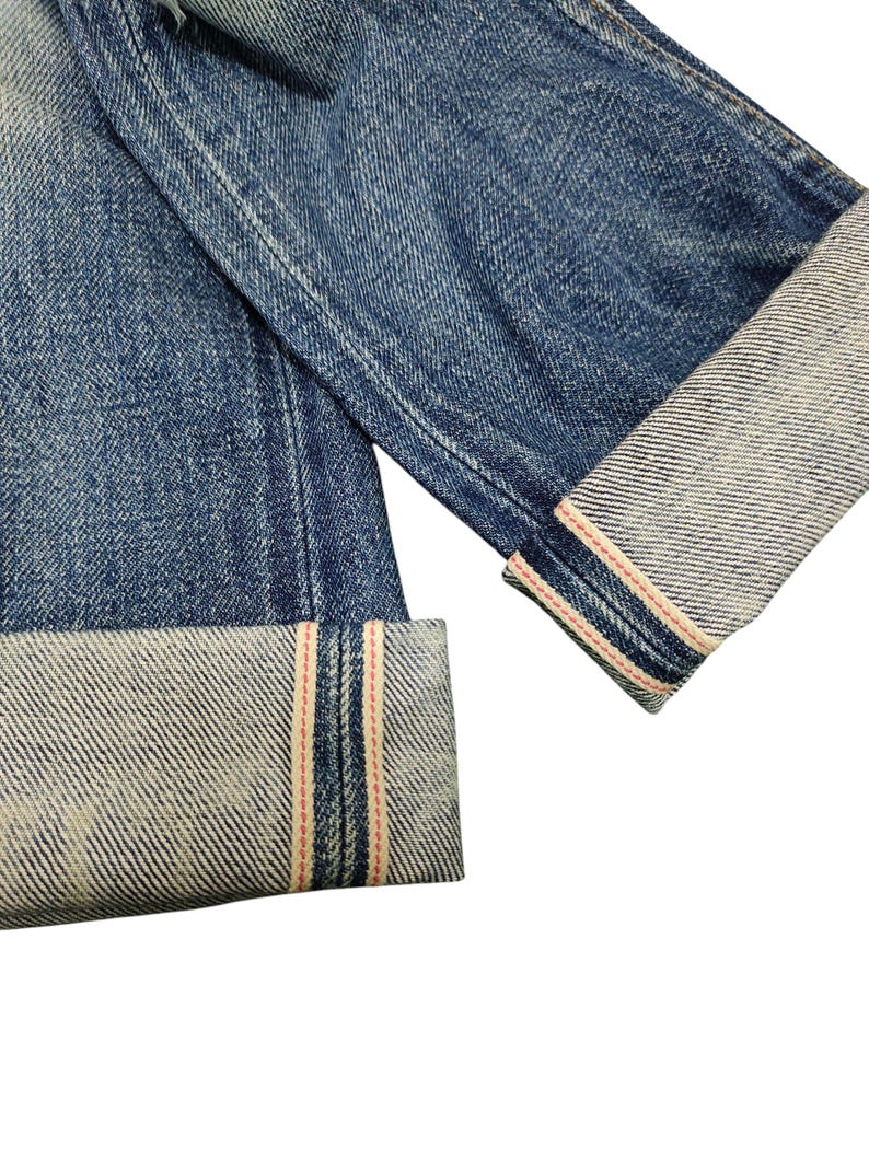 Puede incluir: Primer plano de vaqueros azules con pu&ntilde;os doblados. Los pu&ntilde;os revelan una tela m&aacute;s clara con rayas rojas y blancas a lo largo de los bordes. El denim tiene un aspecto desgastado y texturizado.