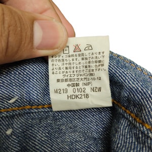 May include: A white care label with black and red text and symbols. The label includes washing instructions, fabric content, and size information. The text reads: "Indigo dye is used, so it will fade. Wash separately. When wet, friction may cause color transfer to other garments. Please be careful. VF Japan (stock) Tokyo Minato-ku Shiba-daimon 2-10-12 Made in China (NIP) M219 0102 NZW HDK218"