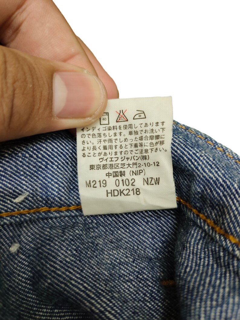 May include: A white care label with black and red text and symbols. The label includes washing instructions, fabric content, and size information. The text reads: "Indigo dye is used, so it will fade. Wash separately. When wet, friction may cause color transfer to other garments. Please be careful. VF Japan (stock) Tokyo Minato-ku Shiba-daimon 2-10-12 Made in China (NIP) M219 0102 NZW HDK218"