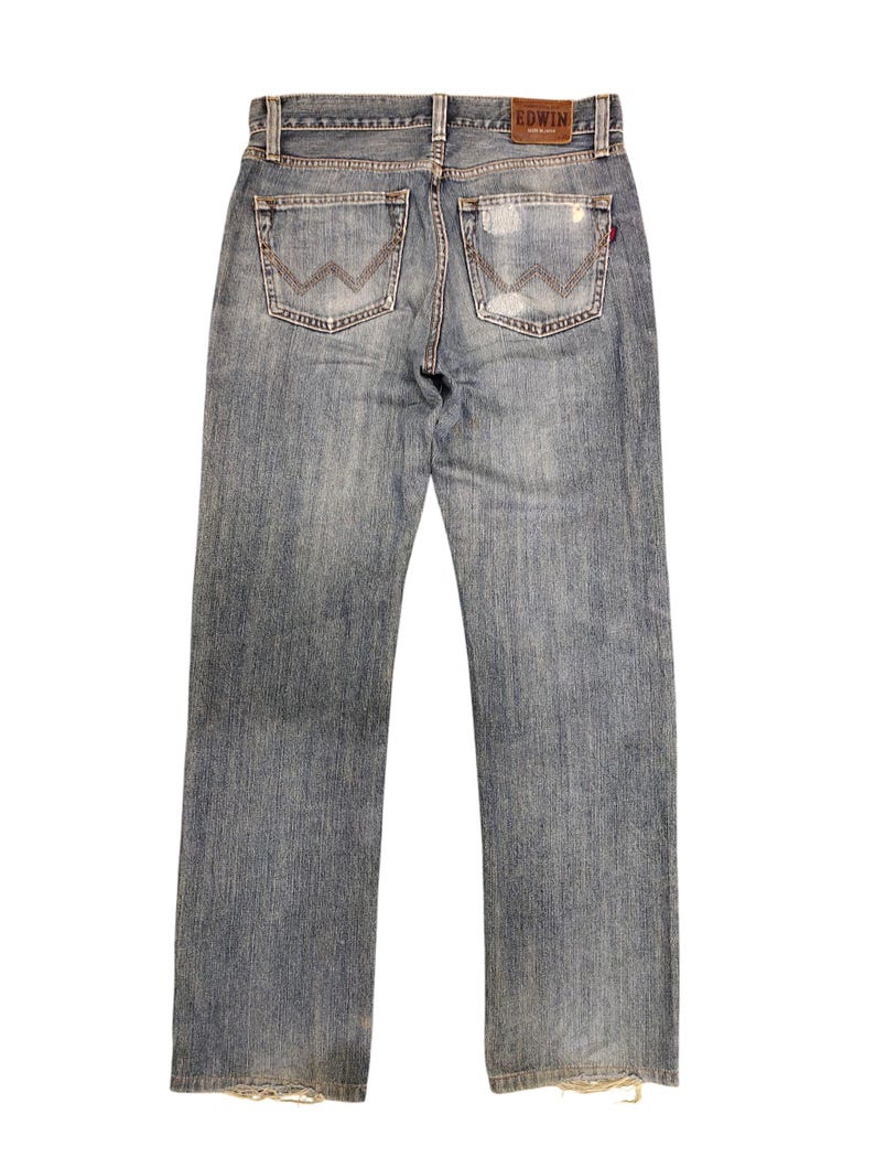 May include: Faded blue denim jeans with a distressed look. Features include two back pockets with a distinctive stitched design, a leather patch with the brand name "EDWIN", and a red tag. The jeans have a straight leg cut and frayed hems.