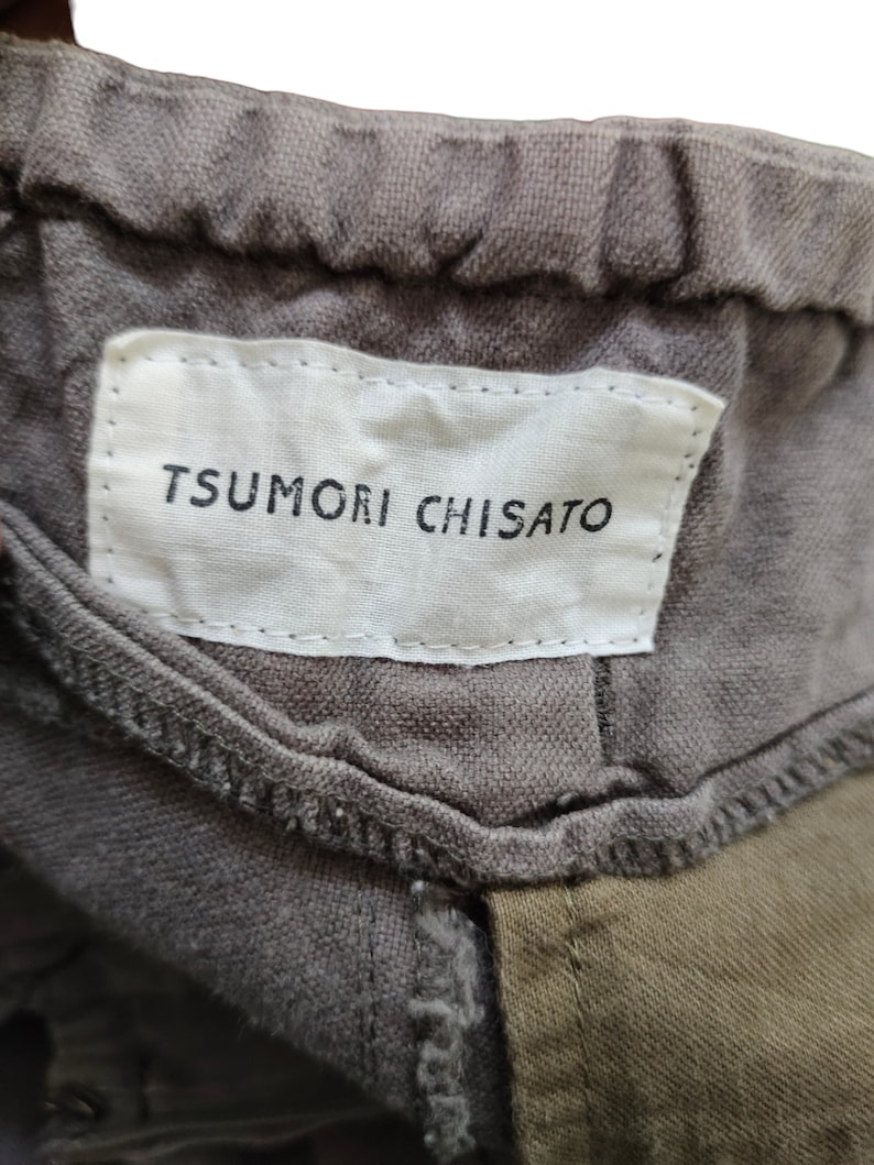 May include: A close-up of a gray fabric label with the text "TSUMORI CHISATO" in black lettering. The label is attached to a garment.