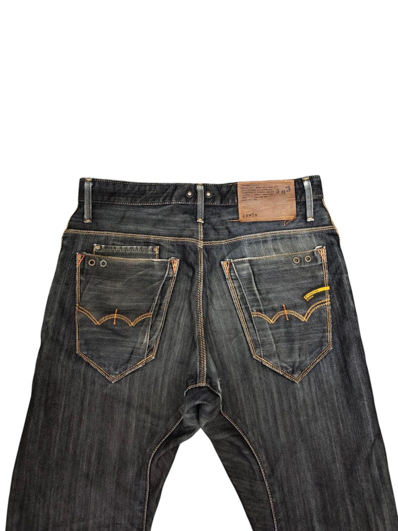Op de afbeelding: Donkerblauwe denim jeans met oranje stiksels en een leren patch. De jeans heeft twee achterzakken met decoratieve stiksels en een gele accent. De merknaam Edwin is zichtbaar op de leren patch.