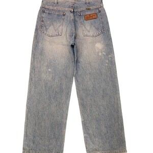 May include: Light blue denim Wrangler jeans with a faded wash. The jeans have a wide leg cut, two back pockets, and a brown leather Wrangler patch. The jeans are a vintage style.