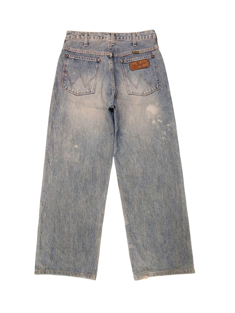 May include: Light blue denim Wrangler jeans with a faded wash. The jeans have a wide leg cut, two back pockets, and a brown leather Wrangler patch. The jeans are a vintage style.