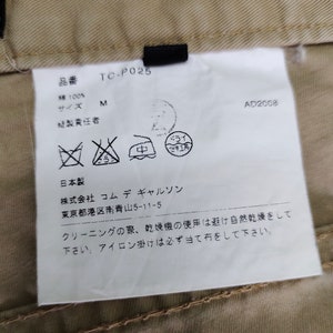 Puede incluir: Una etiqueta blanca con texto negro y s&iacute;mbolos. La etiqueta tiene el texto "TC-P025", "M", "AD2008", "綿100%", "日本製", "株式会社 コムデギャルソン", "東京都港区南青山5-11-5", "クリーニングの際、乾燥機の使用は避け自然乾燥をして下さい。アイロン掛けは必ず当て布をして下さい."