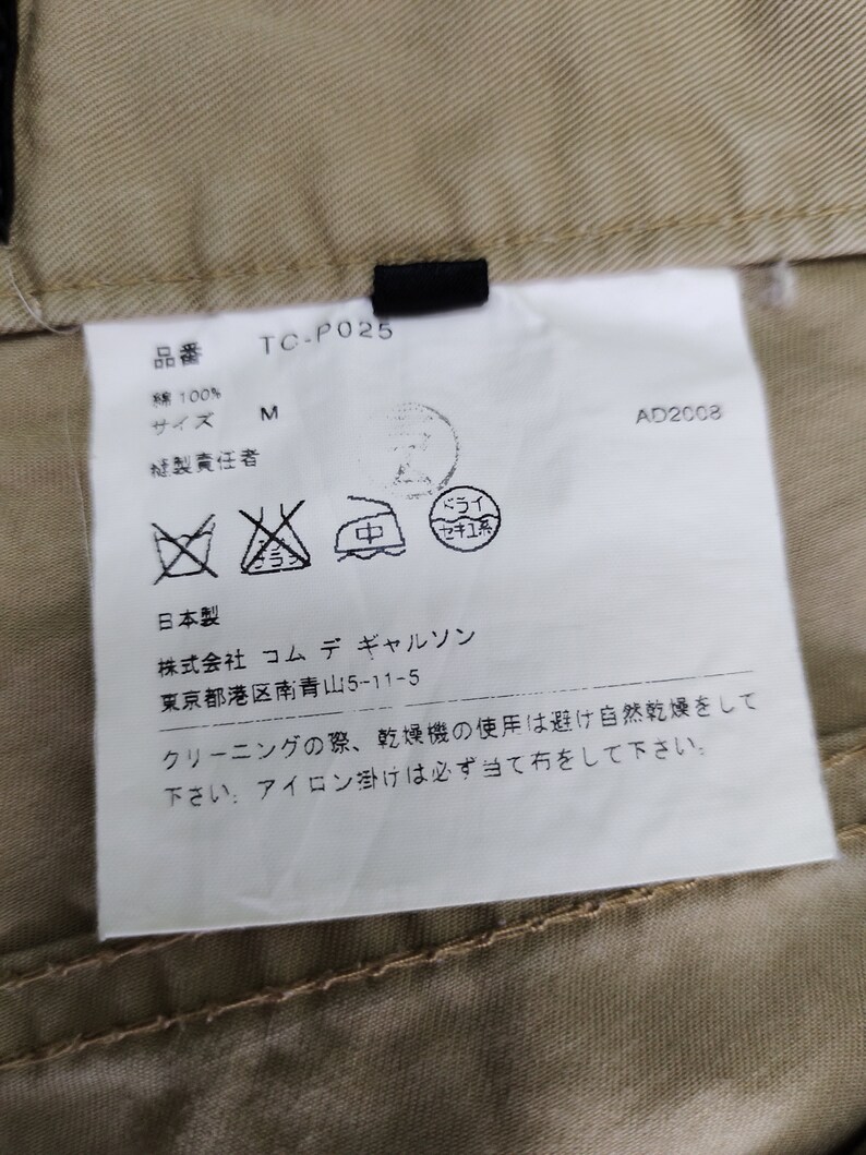 Puede incluir: Una etiqueta blanca con texto negro y s&iacute;mbolos. La etiqueta tiene el texto "TC-P025", "M", "AD2008", "綿100%", "日本製", "株式会社 コムデギャルソン", "東京都港区南青山5-11-5", "クリーニングの際、乾燥機の使用は避け自然乾燥をして下さい。アイロン掛けは必ず当て布をして下さい."