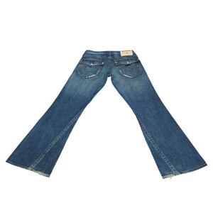 K&ouml;nnte beinhalten: Eine blaue Jeans mit Bootcut-Beinen. Die Jeans hat einen Used-Look und ein kleines rot-wei&szlig;es Label auf der Ges&auml;&szlig;tasche mit dem Schriftzug "True Religion" in Wei&szlig;.