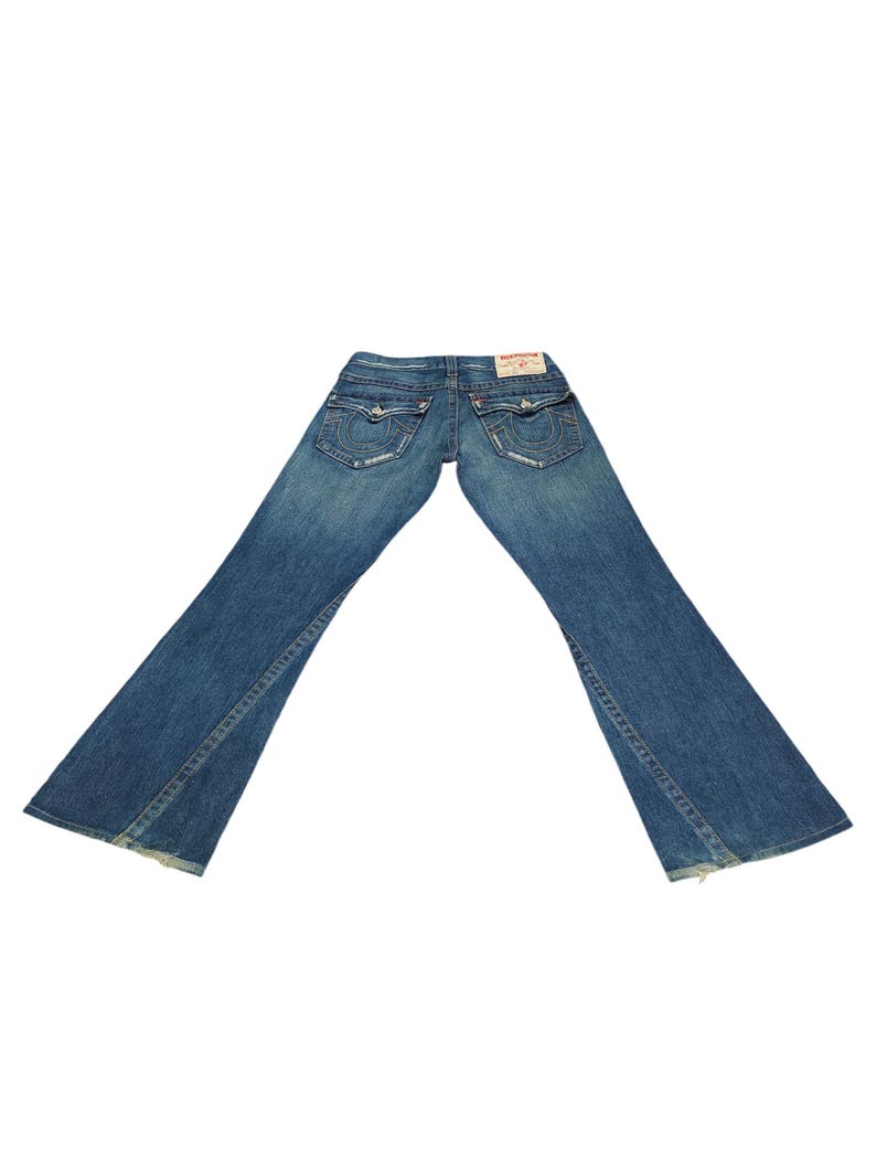K&ouml;nnte beinhalten: Eine blaue Jeans mit Bootcut-Beinen. Die Jeans hat einen Used-Look und ein kleines rot-wei&szlig;es Label auf der Ges&auml;&szlig;tasche mit dem Schriftzug "True Religion" in Wei&szlig;.