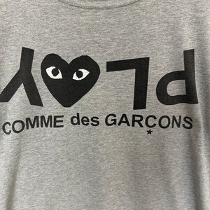 Może przedstawiać: Szara koszulka z czarnym sercem z oczami i nadrukiem "PLAY COMME des GARCONS*" z przodu.