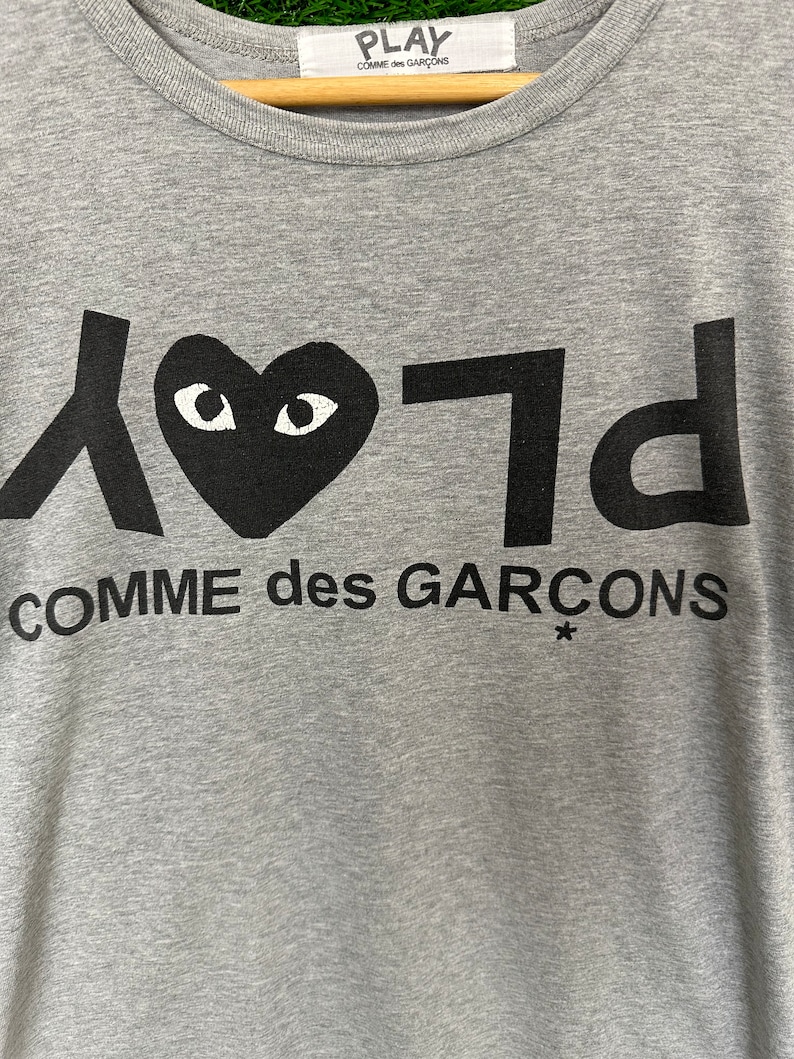 Może przedstawiać: Szara koszulka z czarnym sercem z oczami i nadrukiem "PLAY COMME des GARCONS*" z przodu.