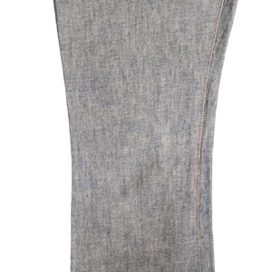 May include: Close-up of the denim fabric of a pair of blue jeans. The fabric has a light blue color and a slightly textured surface. The seam of the jeans is visible along the right edge of the image.