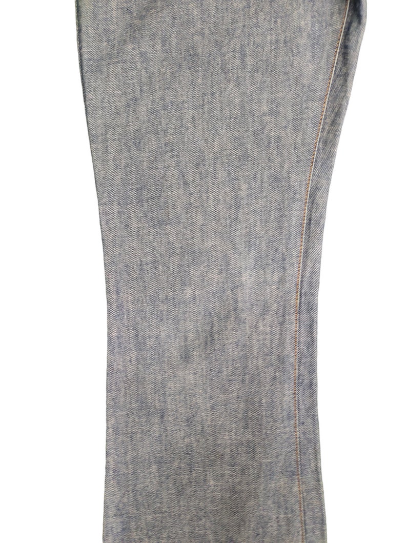 May include: Close-up of the denim fabric of a pair of blue jeans. The fabric has a light blue color and a slightly textured surface. The seam of the jeans is visible along the right edge of the image.