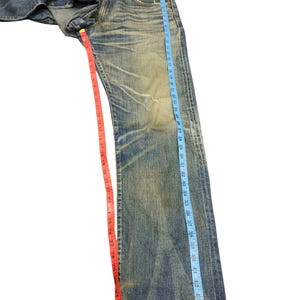 Puede incluir: Vaqueros de mezclilla azul descolorido con un aspecto desgastado. Los vaqueros se miden con una cinta m&eacute;trica azul que muestra una longitud de aproximadamente 117 cm. Una cinta m&eacute;trica roja muestra una longitud de aproximadamente 84 cm.