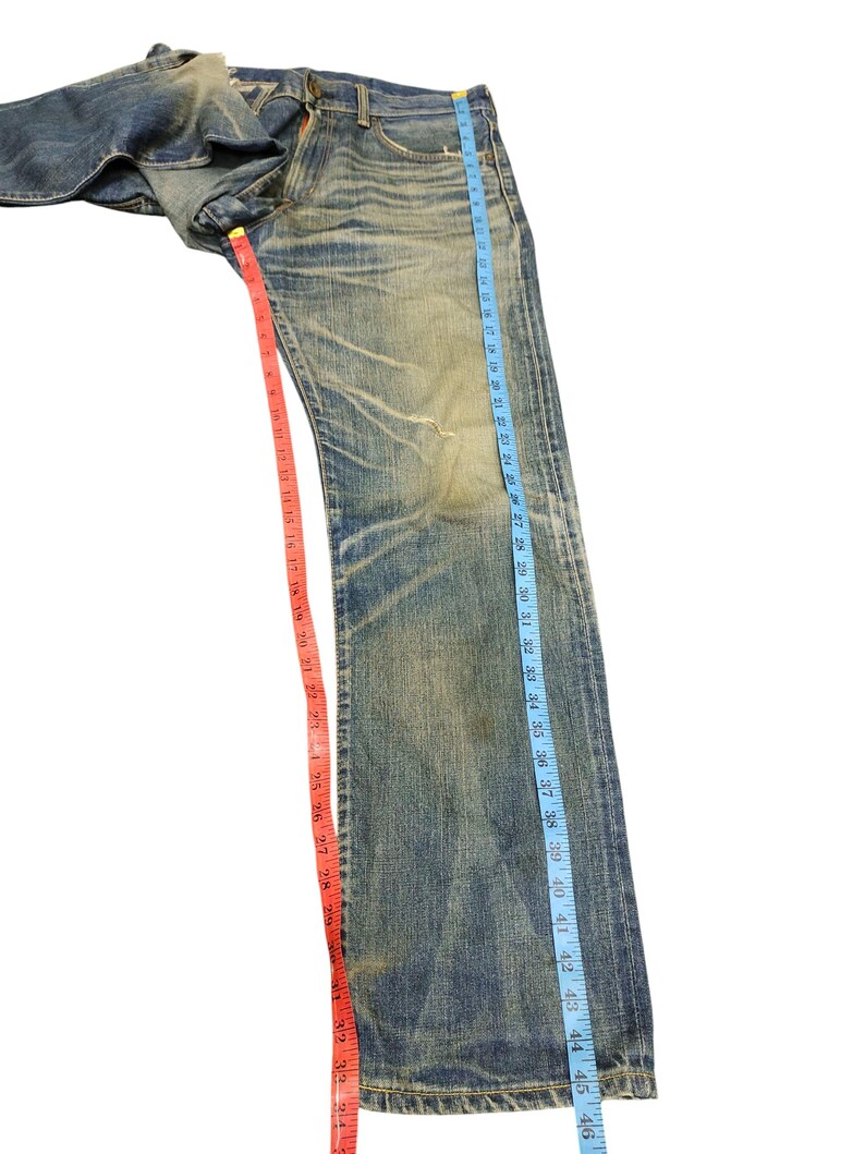Puede incluir: Vaqueros de mezclilla azul descolorido con un aspecto desgastado. Los vaqueros se miden con una cinta m&eacute;trica azul que muestra una longitud de aproximadamente 117 cm. Una cinta m&eacute;trica roja muestra una longitud de aproximadamente 84 cm.