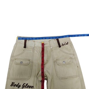 Puede incluir: Pantalones vaqueros de color caqui con costuras y detalles marrones. Cuenta con dos bolsillos delanteros, cierre de bot&oacute;n y el texto "Body Glove South Bay" bordado en la parte delantera. La palabra "SoCal" est&aacute; bordada en la parte trasera. Una cinta m&eacute;trica est&aacute; colocada sobre los pantalones.